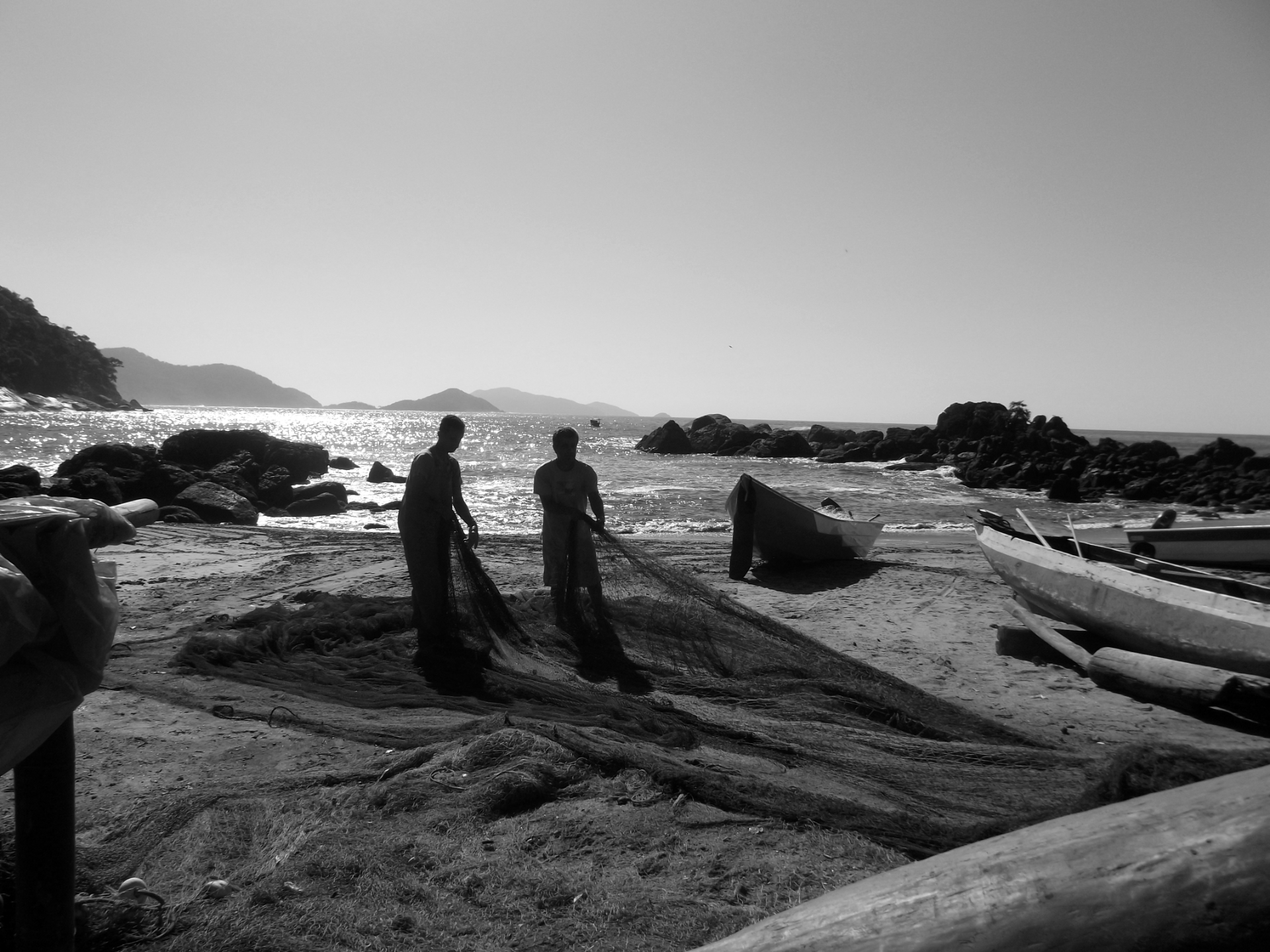 Foto em preto e branco. No centro da imagem dois homens estão puxando e esticando uma grande rede de pesca sobre a areia da praia. Atrás deles há uma canoa e dois barcos. Ao fundo o mar está com ondas leves, com sol forte e o céu sem nuvens. 
