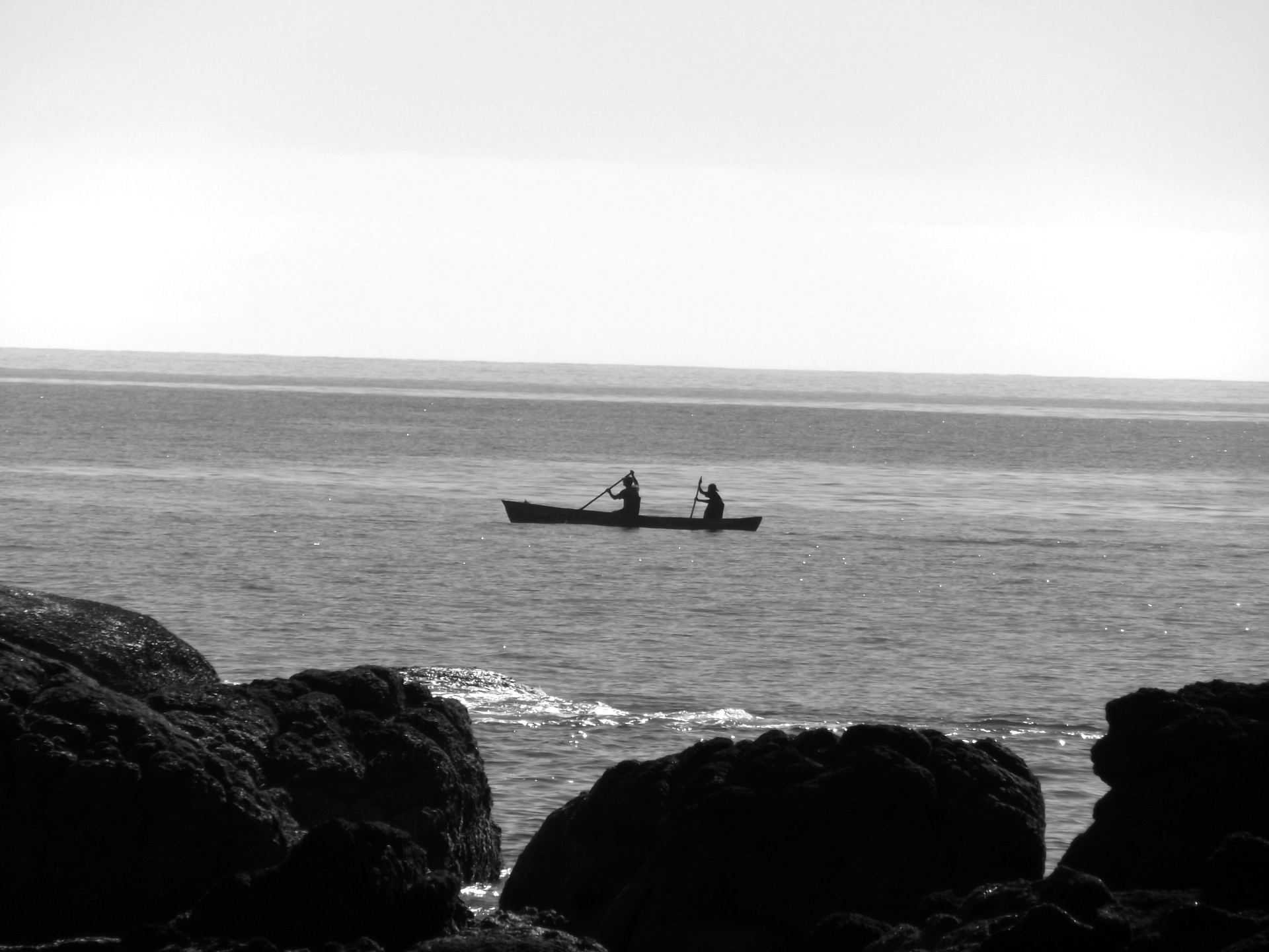 No terço superior o céu está limpo. Ao centro vê-se a silhueta de duas pessoas remando uma canoa no mar. A água está calma, com poucas ondas. No terço inferior há uma formação rochosa.